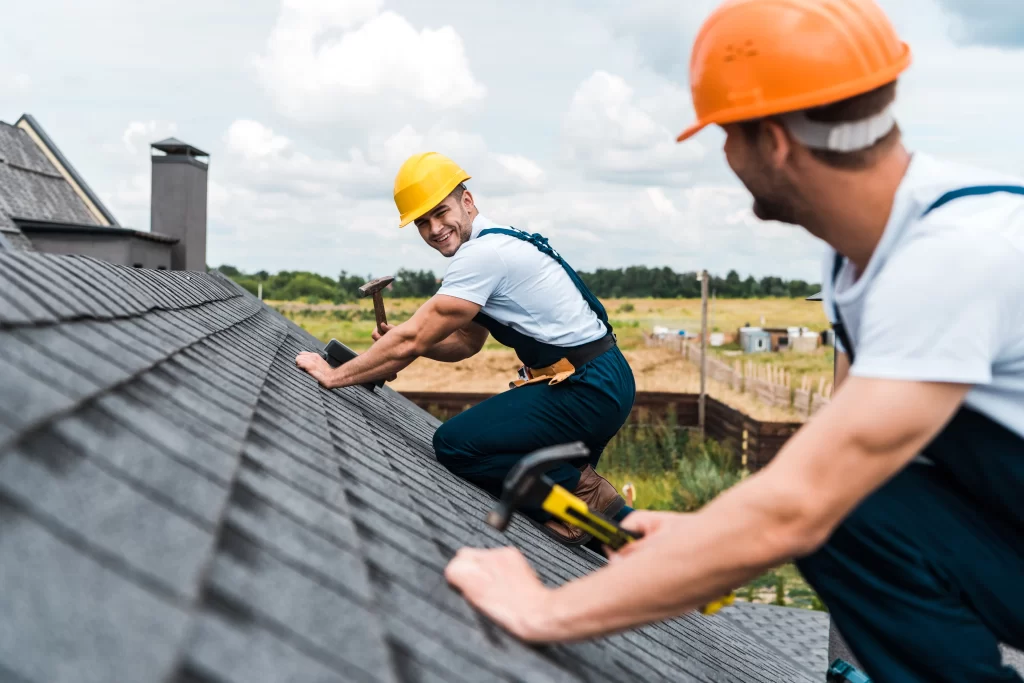 Two uniformed carpenters smiling at each other while fixing the roof of the client.