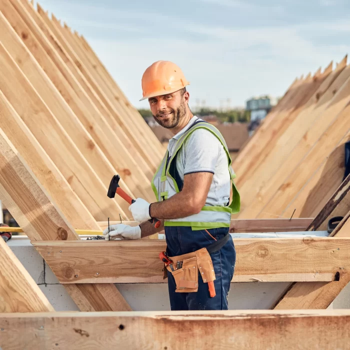 A builder smiling while hammering a nail on the upside down roof wooden frame.